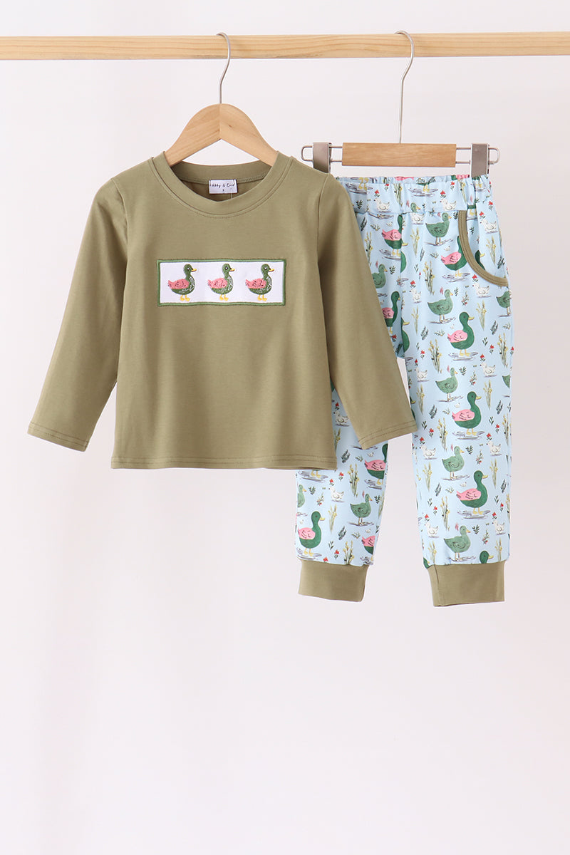 A children's two-piece pajama set hanging on wooden hangers against a plain light background. The set includes an olive green long-sleeve top with a rectangular duck-print patch on the chest, and light blue patterned pants with green ducks and pink watermelon motifs.