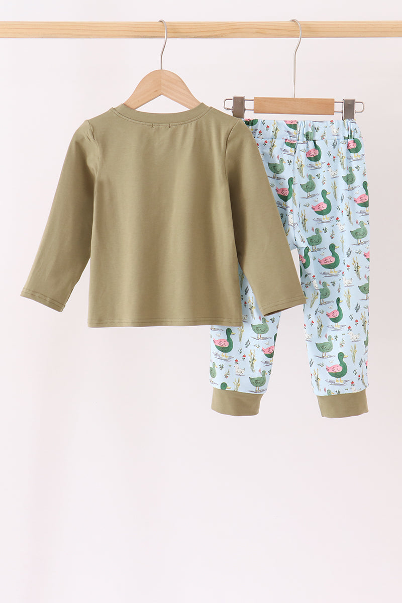 A two-piece children's pajama set hanging on wooden hangers from a light wood rod. The set includes a solid olive green long-sleeve top and matching green cuffs, paired with light blue pants covered in a green cactus and pink watermelon print.