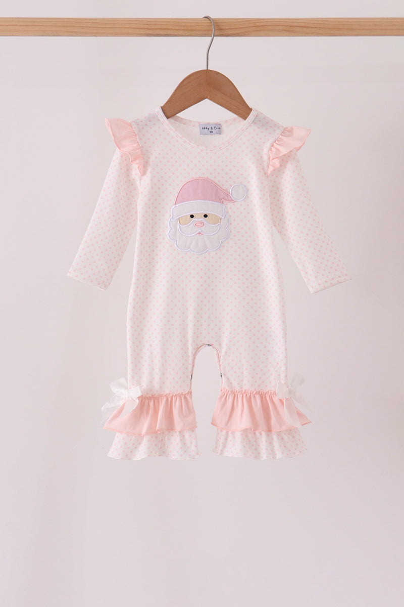 A baby long-sleeve romper in white with tiny pink polka dots, featuring a cute embroidered Santa face on the chest, pink ruffle details on the shoulders and legs, and white bows at the ankle cuffs. The romper is hanging on a wooden hanger from a light wood rod against a plain off-white background.