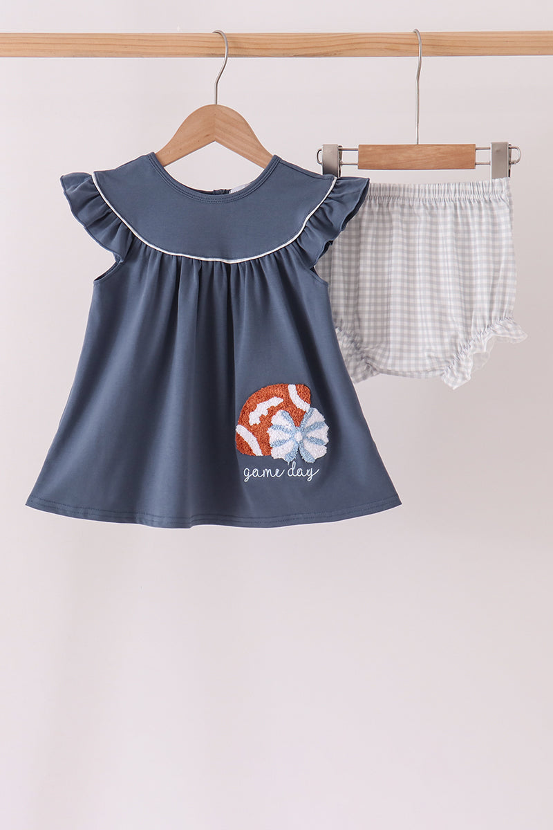 A toddler girl's two-piece outfit hanging on a wooden hanger bar. The set includes a slate blue sleeveless top with ruffled shoulders and a 'game day' graphic, paired with white and gray gingham check shorts with ruffled trim.