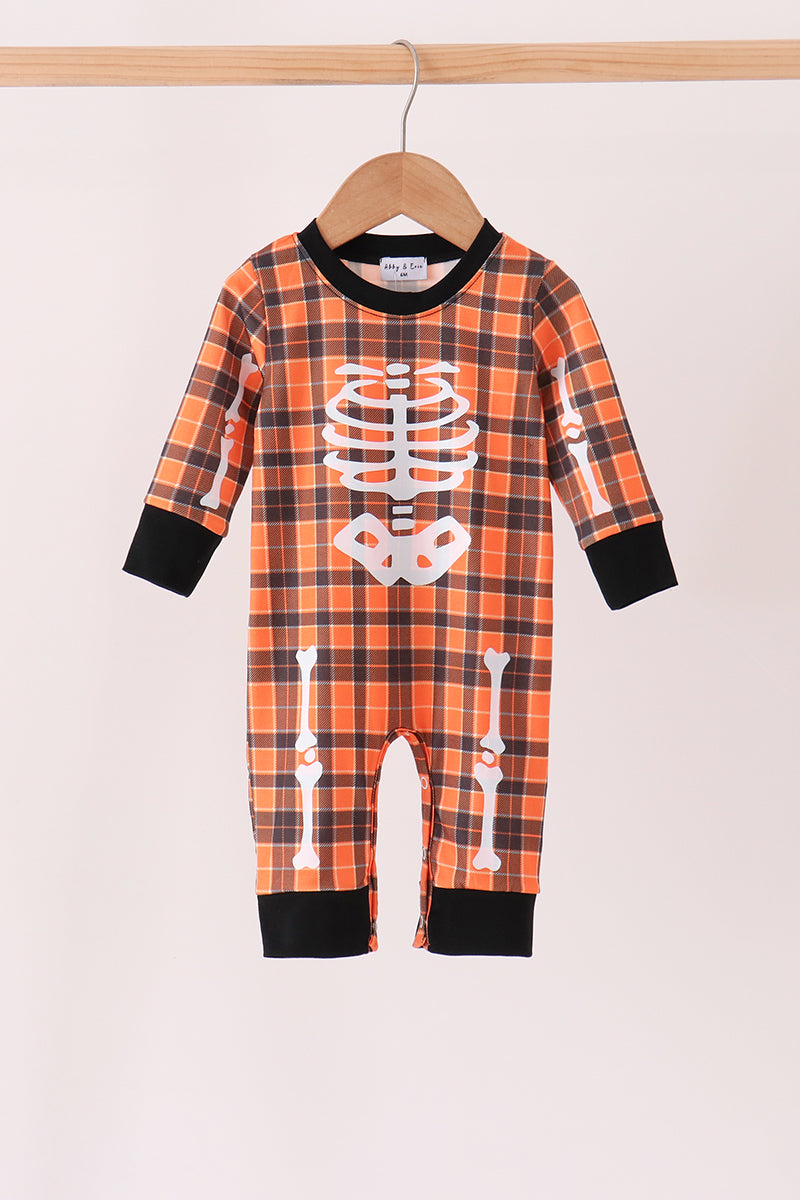 A baby skeleton-themed plaid jumpsuit hanging on a wooden hanger. The jumpsuit features an orange and black plaid pattern with white skeleton rib cage and leg bone graphics, black ribbed collar, cuffs, and hems.