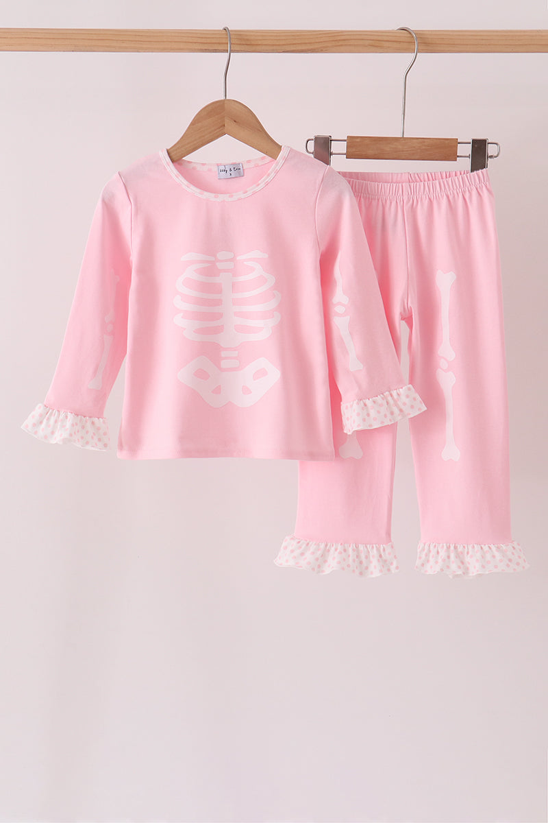 A two-piece children's pajama set in light pink, featuring a long-sleeve top with a white skeleton rib cage graphic and white ruffled cuffs, paired with matching pants with white ruffled hems. The set is displayed on wooden hangers against a light pink background.