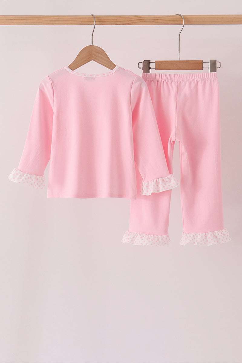 A two-piece children's pajama set in light pink, featuring a long-sleeve top and matching pants, both hanging on wooden hangers from a wooden rod. The set has delicate white lace trim details at the cuffs of the top and the hems of the pants.