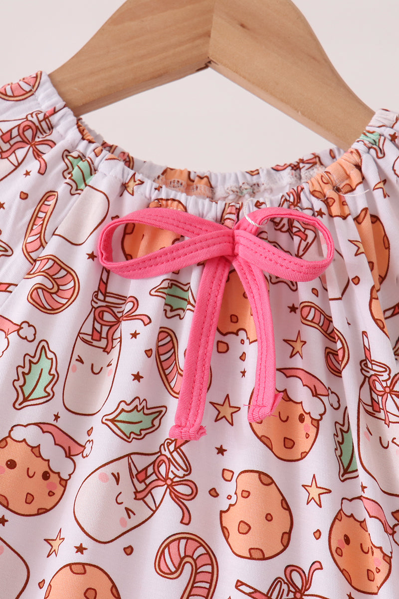 A close-up of a children's top on a wooden hanger, featuring a white base with a festive holiday print of cookies, candy canes, and stockings in orange, green, and red, accented by a bright pink drawstring bow at the neckline.