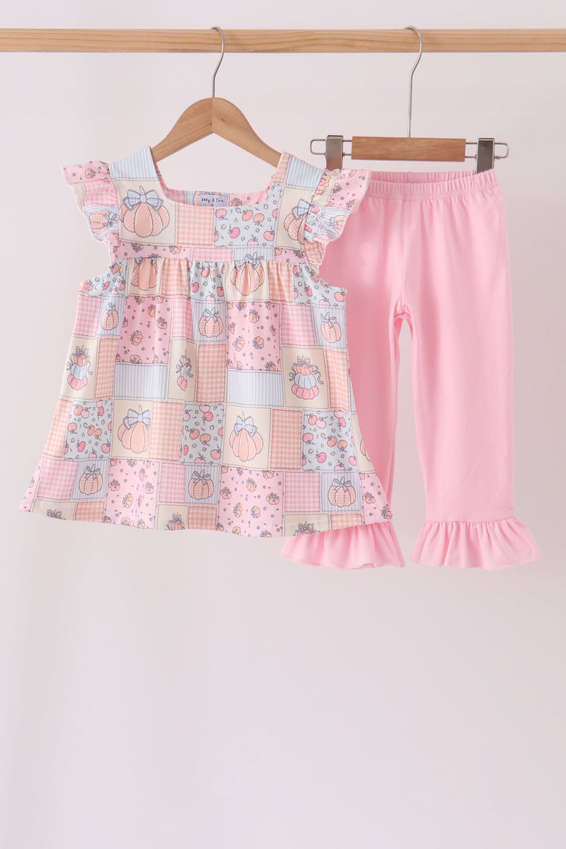 A toddler girl's two-piece clothing set hanging on wooden hangers against a plain light background. The set includes a tiered patchwork dress in pastel pink, peach, and light blue with pumpkin and floral motifs, and matching solid light pink ruffled leggings.