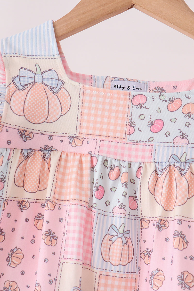 A close-up of a baby dress with a patchwork design featuring pink pumpkins, strawberries, gingham, and striped patterns, hanging on a wooden hanger.