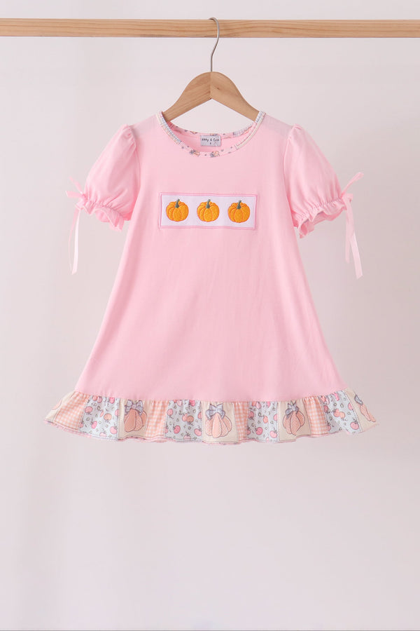 A pink short-sleeve children's dress hanging on a wooden hanger against a light beige wall. The dress has three orange pumpkins on a white patch on the chest, a ruffled hem with a peach and floral print, and ribbon ties on the sleeves.