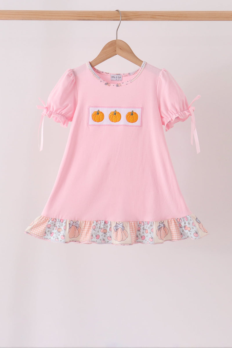 A pink short-sleeve children's dress hanging on a wooden hanger against a light beige wall. The dress has three orange pumpkins on a white patch on the chest, a ruffled hem with a peach and floral print, and ribbon ties on the sleeves.