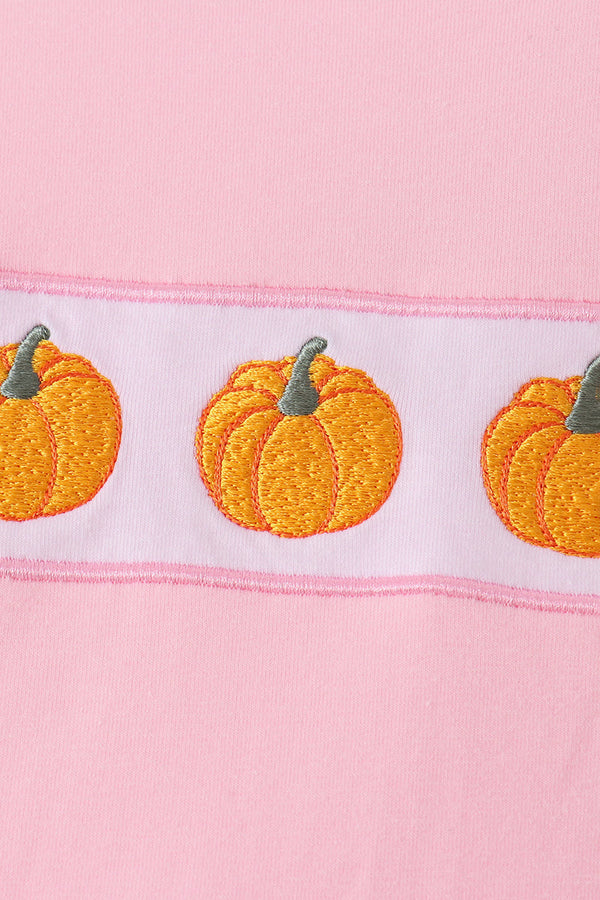 A close-up of a pink fabric item featuring a horizontal white band with embroidered orange pumpkins and green stems.