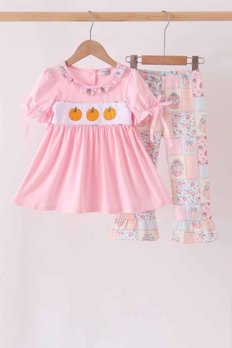 A toddler girl's two-piece clothing set hanging on wooden hangers against a plain light background. The set includes a light pink short-sleeve dress with three orange pumpkins on the chest and matching patchwork-print pants.