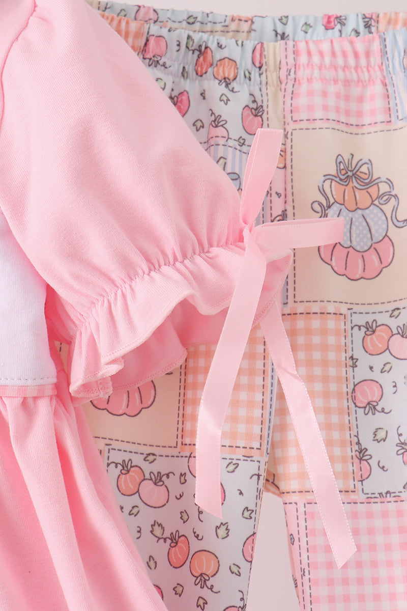 A close-up of a pink children's dress or romper featuring a patchwork-style print with gingham, pumpkin motifs, and a pink ribbon tie at the waist.