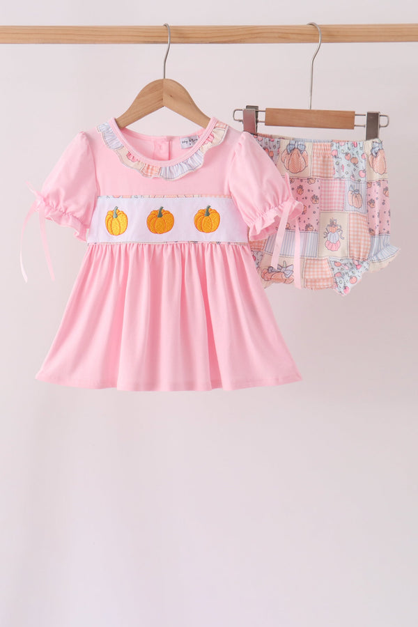 A baby girl's two-piece outfit set hanging on wooden hangers against a plain light background. The set includes a light pink short-sleeve dress with a white smocked chest panel featuring three orange pumpkins, and matching patchwork-print baby shorts in pastel colors.