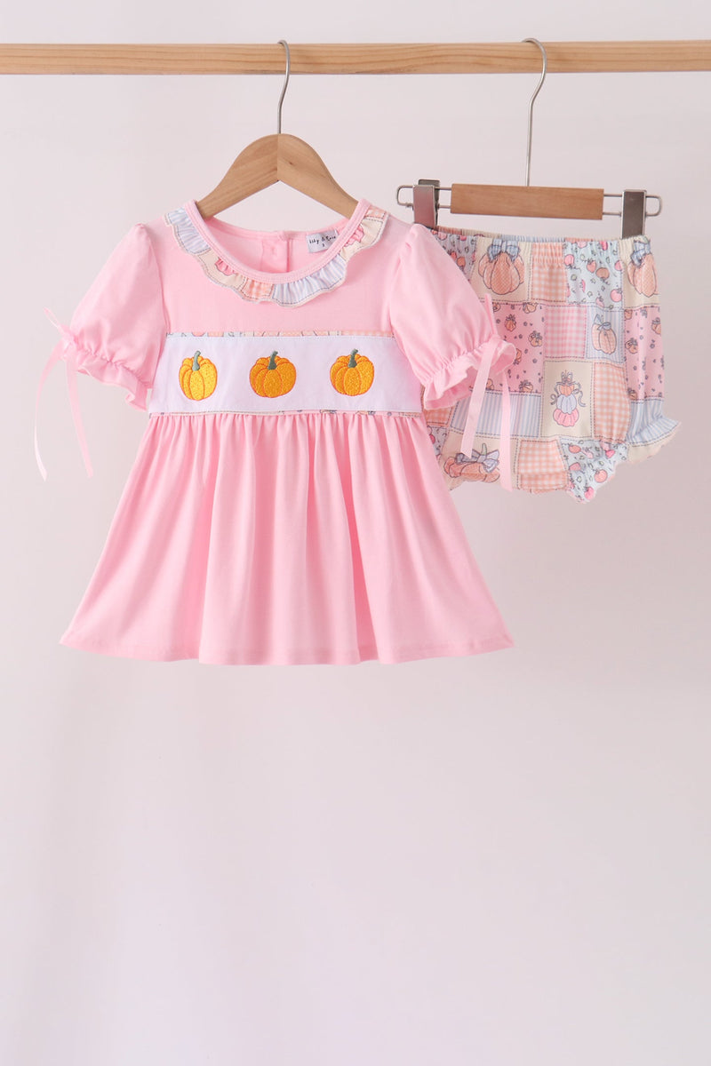 A baby girl's two-piece outfit set hanging on wooden hangers against a plain light background. The set includes a light pink short-sleeve dress with a white smocked chest panel featuring three orange pumpkins, and matching patchwork-print baby shorts in pastel colors.