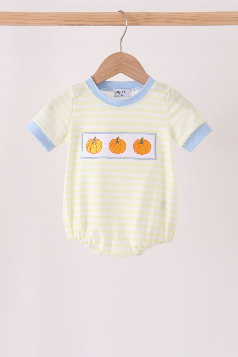 A baby short-sleeve bodysuit with pale yellow and white horizontal stripes, light blue trim at the collar and cuffs, and a chest graphic of three orange pumpkins, hanging on a wooden hanger against a light beige background.