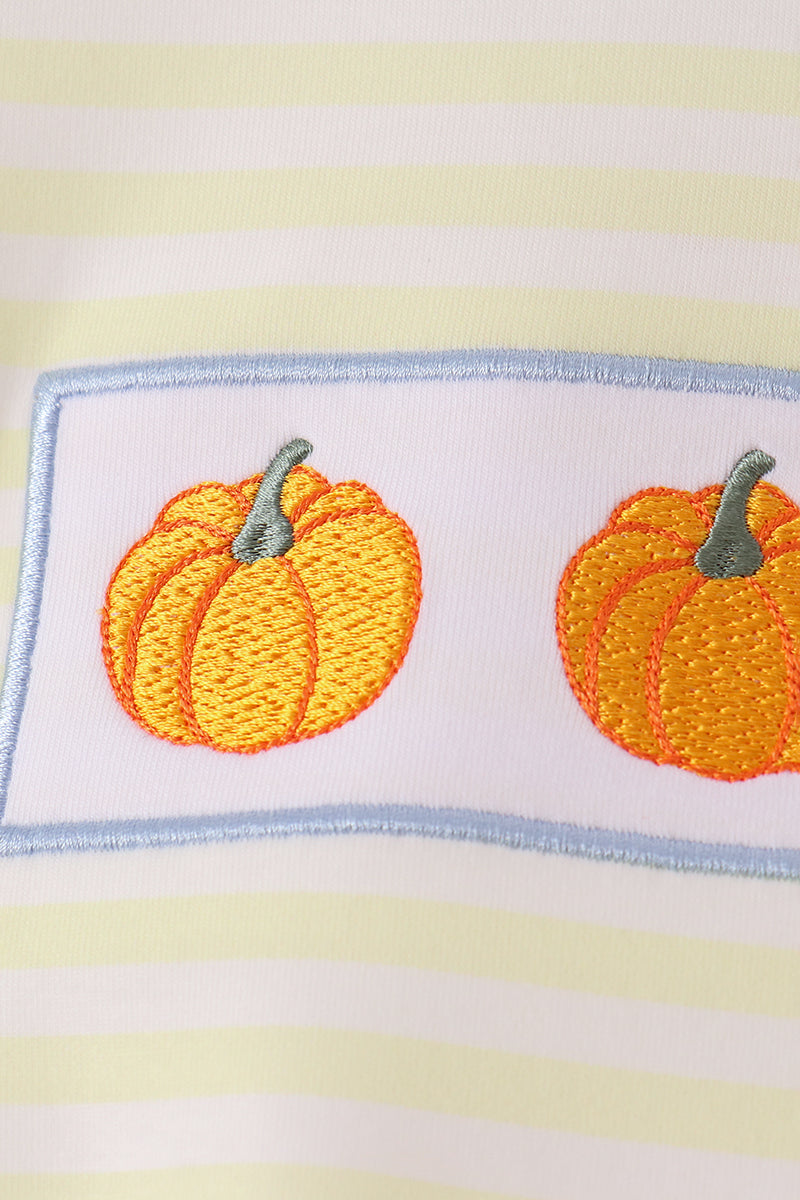 A close-up of a white fabric item featuring two embroidered orange pumpkins with gray stems inside a light blue rectangular border, set against a background of pale yellow and white horizontal stripes.