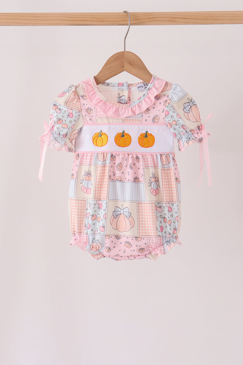 A baby girl's short-sleeve patchwork romper in soft pink, peach, and light blue, featuring three embroidered orange pumpkins across the chest, hanging on a wooden hanger against a light beige background.