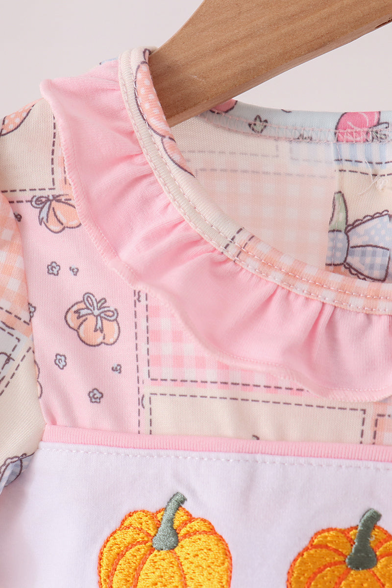 Close-up of a baby dress featuring a pink ruffled collar, a patchwork print with pumpkins and gingham, and a white lower section with embroidered orange pumpkins.