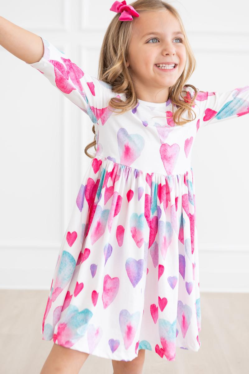 Smiling girl wearing a Light Hearted 3/4 Sleeve Twirl Dress with Pockets for Girls, showcasing a colorful heart pattern. Perfect for play and special occasions, this 3/4 sleeve twirl dress allows for easy movement and twirling.