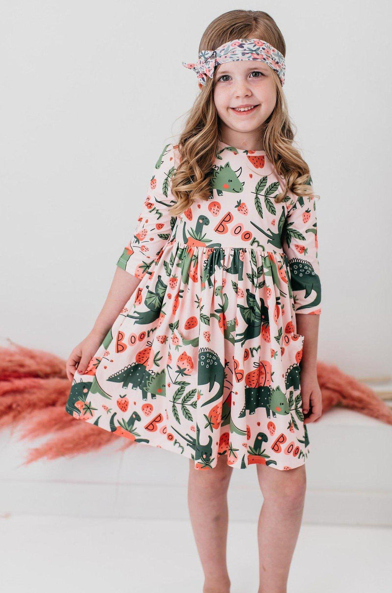 'Hear Me Roar' 3/4 Sleeve Pocket Twirl Dress for Girls worn by a cheerful girl posing with hands on hips, showcasing its fun dinosaur design with strawberries and the word 'Boom'.