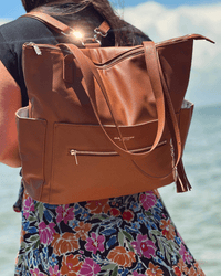 Woman wearing cognac convertible diaper bag as backpack near water, showing adjustable straps and textured exterior in