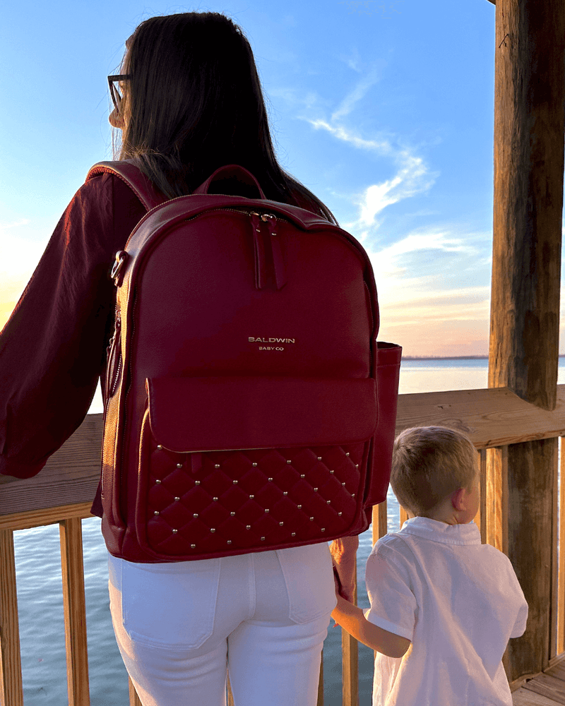 Crimson vegan leather diaper bag with quilted front pocket worn by mother and child overlooking coastal water at sunset.