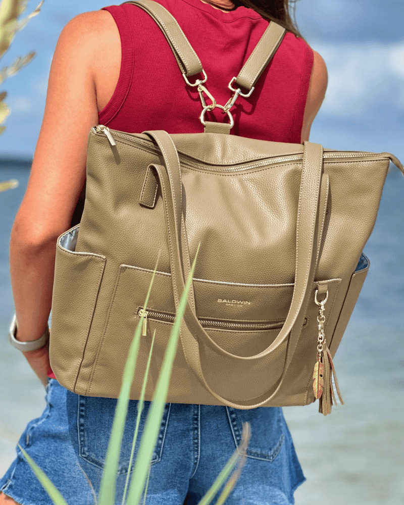 Woman wearing taupe convertible diaper bag as backpack with gold hardware against beach background