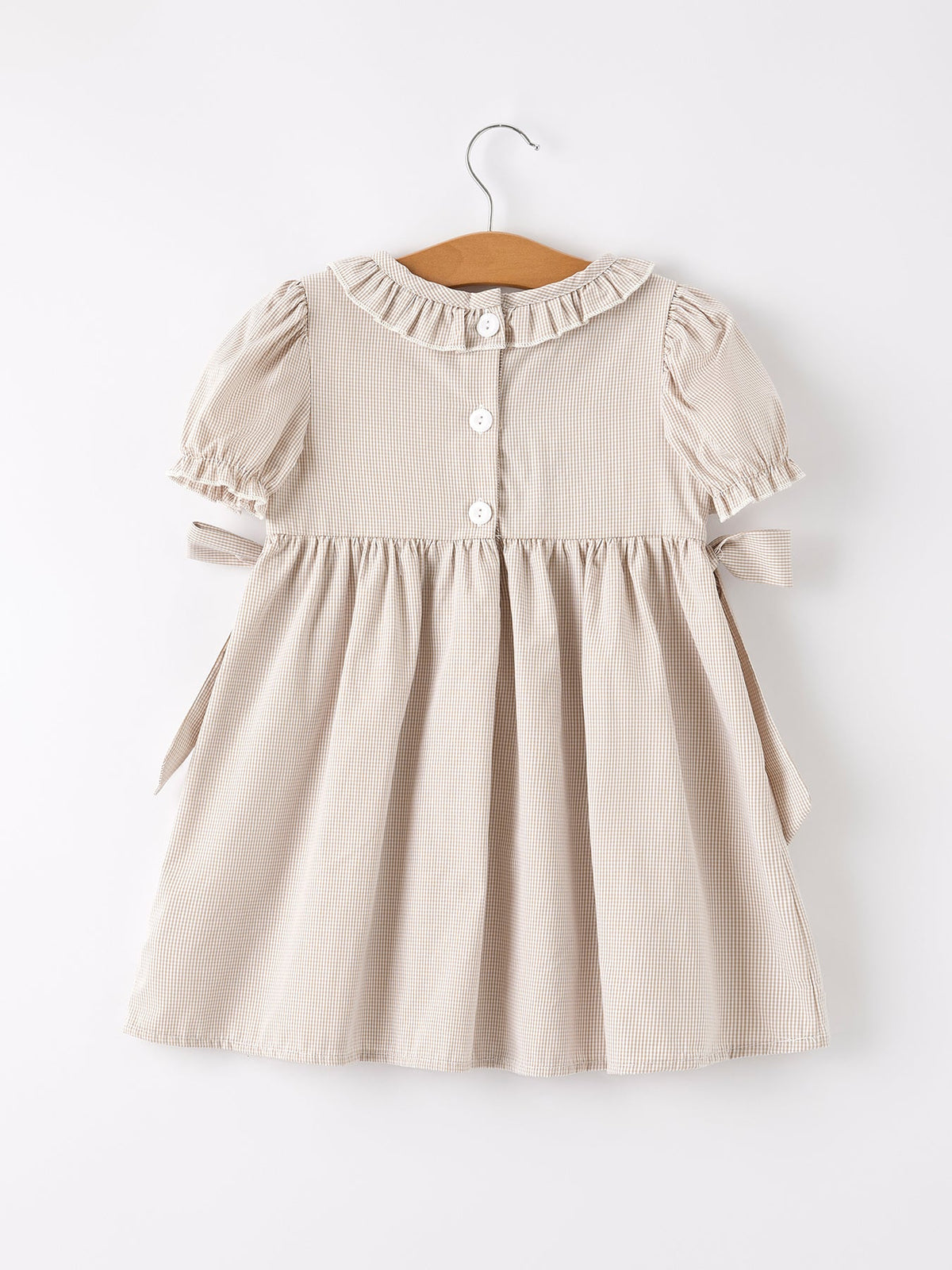 Beige plaid girls' smock dress with a ruffled collar, white buttons, puff sleeves, and side waist bows on a hanger.