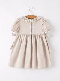 Beige plaid girls' smock dress with a ruffled collar, white buttons, puff sleeves, and side waist bows on a hanger.