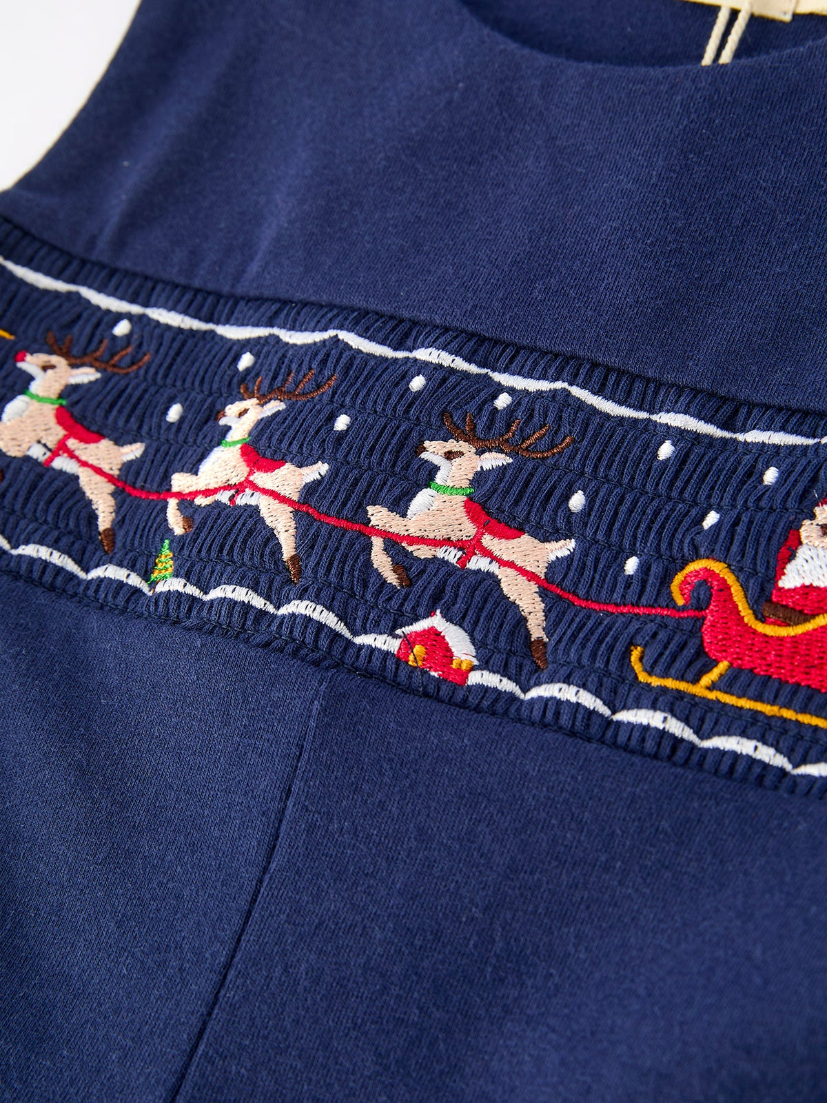 Navy baby romper featuring embroidered reindeer pulling Santa's sleigh on a smocked waistband with festive snow details.