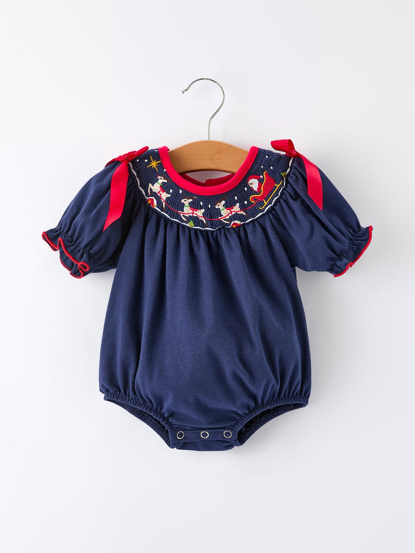 Navy baby romper with embroidered Santa sleigh and reindeer smock, red ribbon shoulder ties, and puffed sleeves. Festive Christmas outfit.