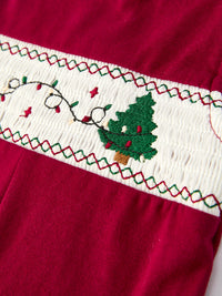 Red baby romper featuring white smocking with embroidered Christmas tree, string lights, and festive red and green zigzag stitching details.
