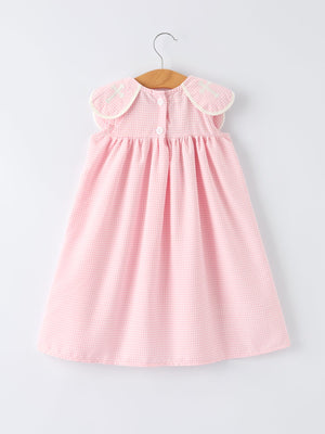 Pink gingham christening dress for girls with white-trimmed shoulder patches featuring cross embroidery, on hanger.