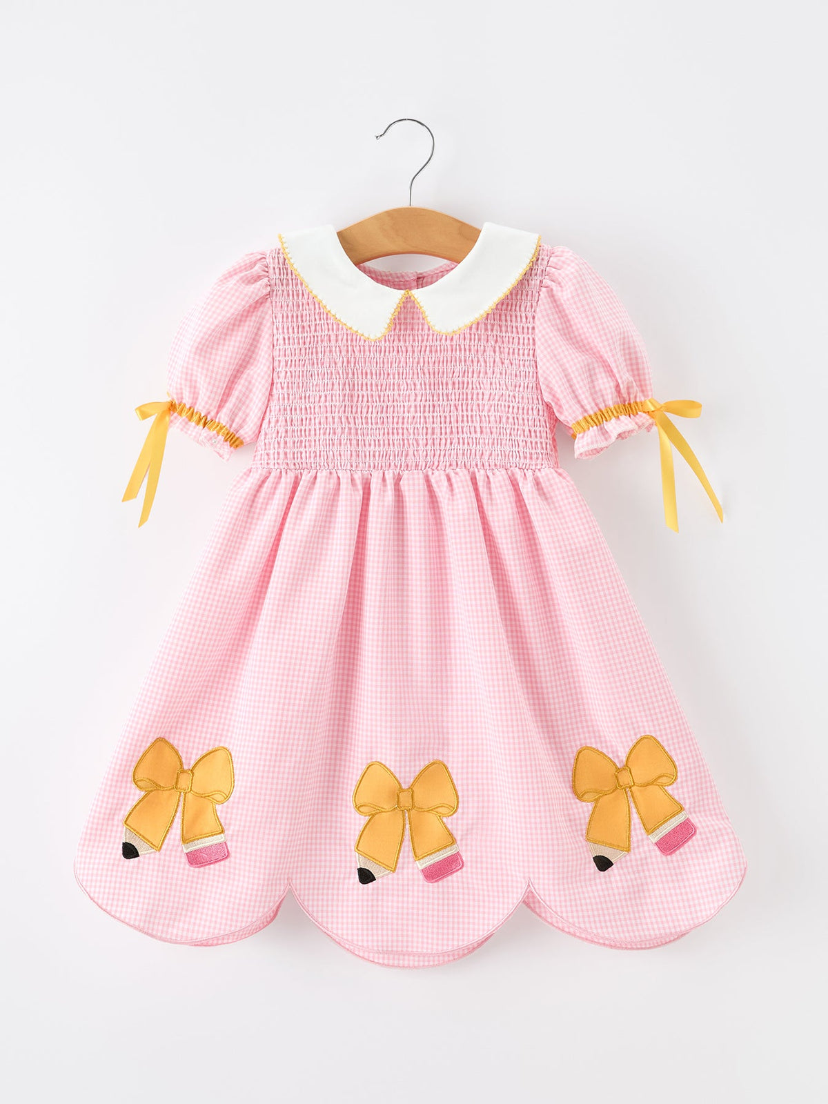 Displaying the Girls Back To School Pencil Twirl Dress with bow appliques, featuring a pink gingham design hanging on a hanger.