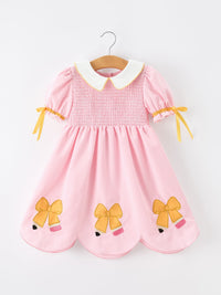 Displaying the Girls Back To School Pencil Twirl Dress with bow appliques, featuring a pink gingham design hanging on a hanger.