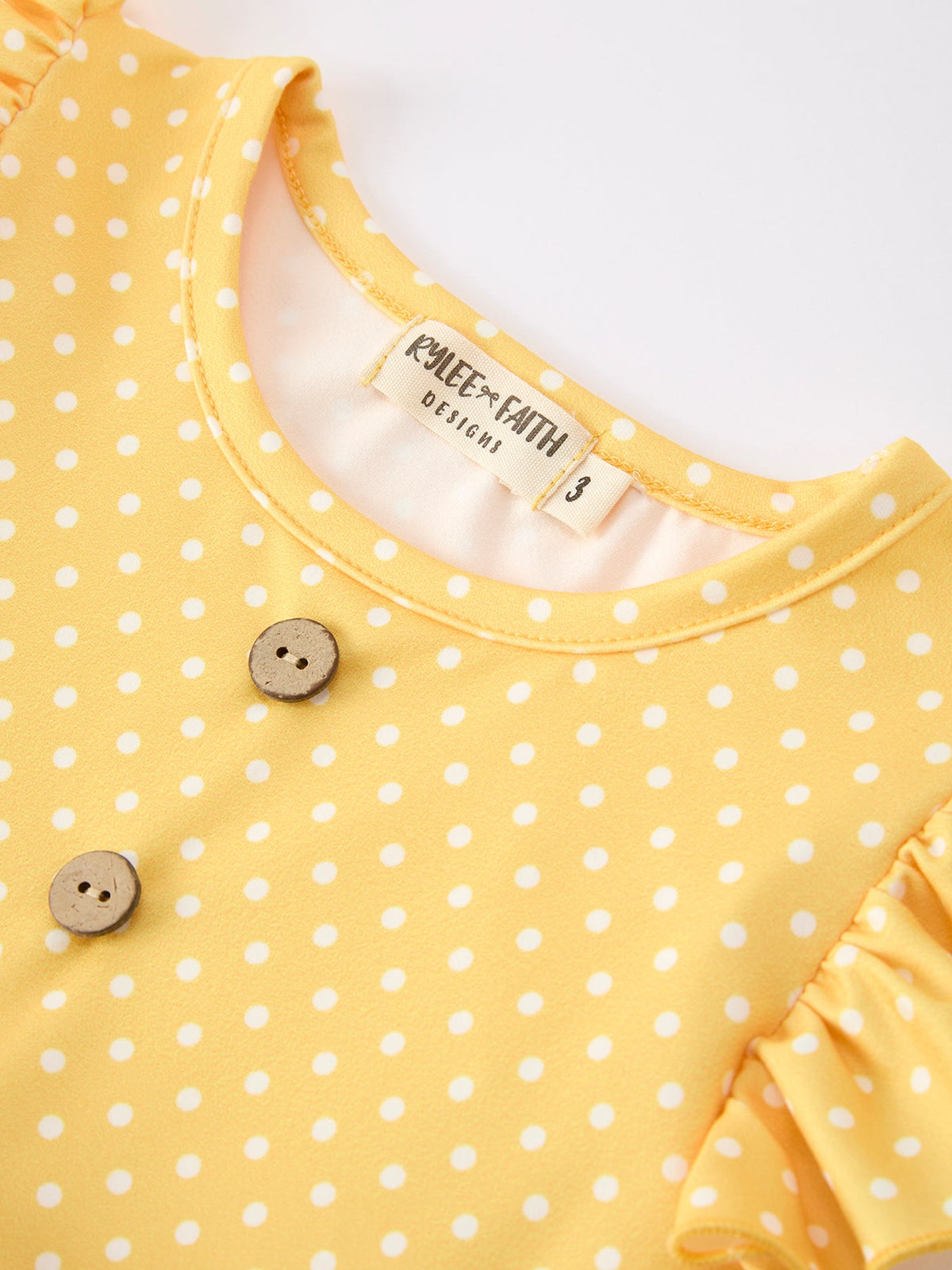 Close-up of the neckline from the Girls Back To School Rainbow School Bus Ruffle Dress Leggings Set, featuring a soft yellow fabric with polka dots and wooden buttons.