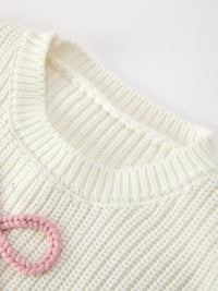 Close-up of a cream-colored ribbed knit sweater with a pink braided ring detail on the chest, set against a plain light background.
