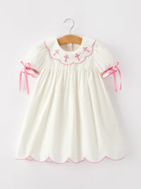Ivory girl's baptism dress with pink cross embroidery on scalloped collar, puff sleeves with ribbon ties, and scalloped hem.