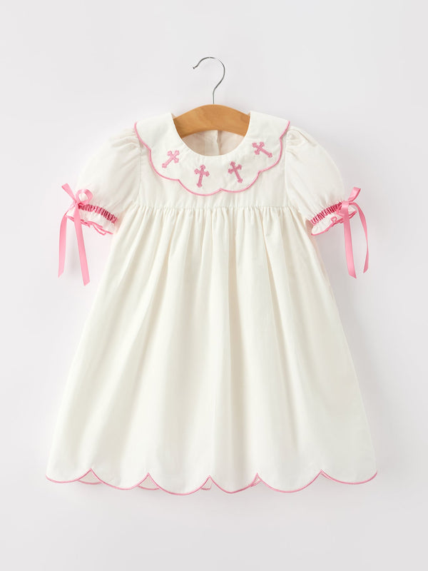 Ivory girl's baptism dress with pink cross embroidery on scalloped collar, puff sleeves with ribbon ties, and scalloped hem.