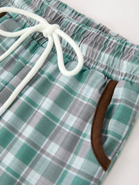 Close-up of green plaid pants waistband showing gathered elastic fabric, white rope drawstring bow, and brown piping pocket