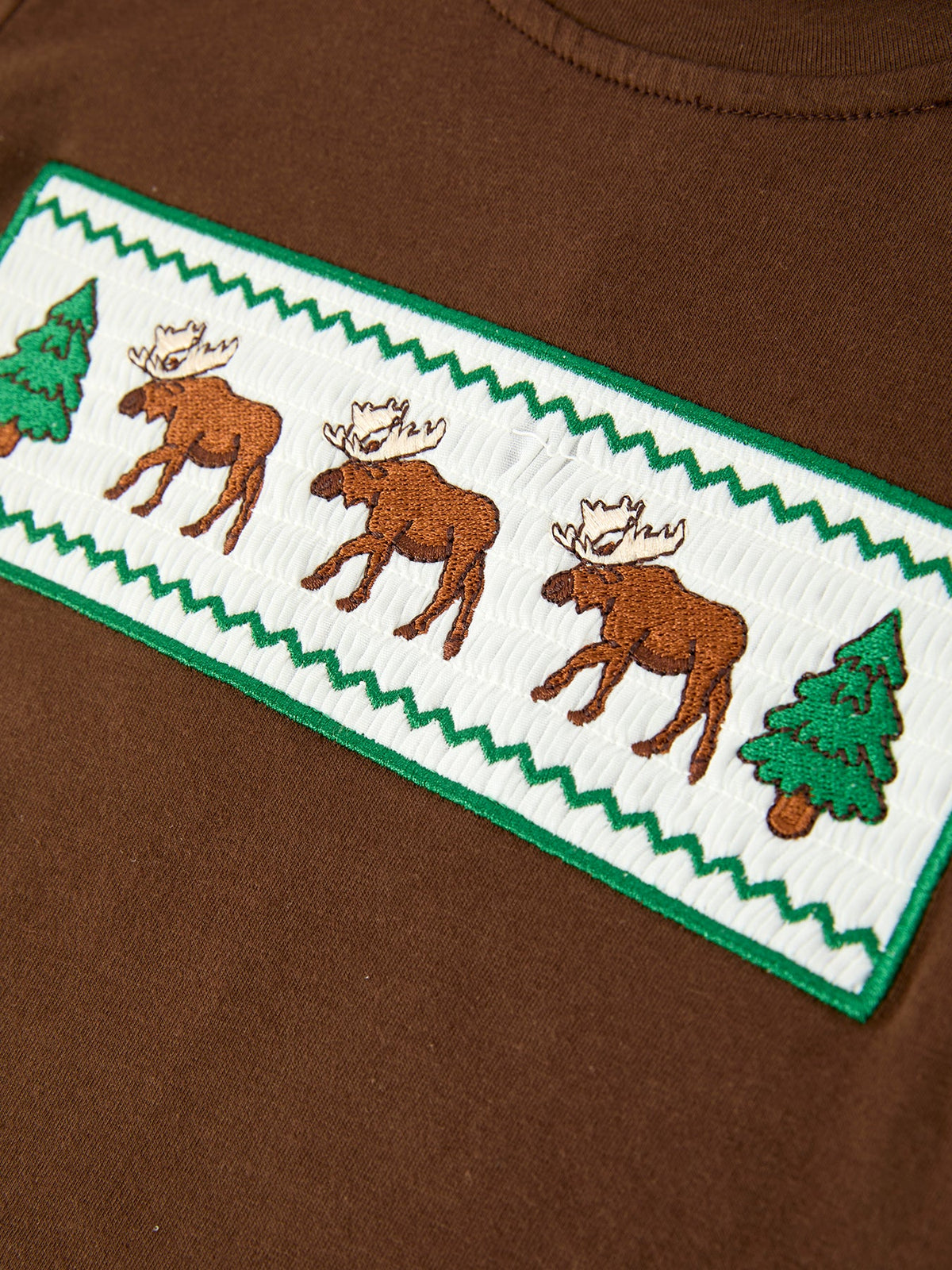 Macro shot of white smocked chest patch with three brown moose, green pine trees, and zigzag border stitching
