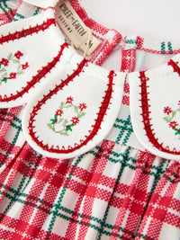 Red and green plaid baby romper with white bib collar featuring embroidered red floral wreath and red stitched trim.