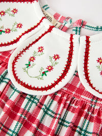 Close-up of red and green plaid baby dress featuring white collar with embroidered holiday wreath and red stitched trim.