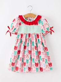 A toddler girl's short-sleeve dress hanging on a wooden hanger against a plain white wall. The dress has a light pink base with a repeating pattern of red bows, teal bows, and teal pine trees, featuring a red ruffled collar, a light teal smocked chest band, and red ribbon ties on the sleeves.