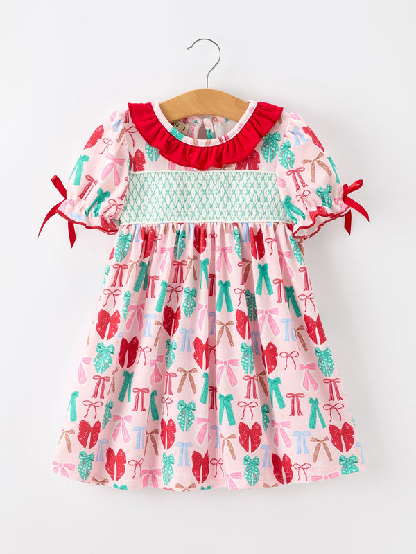 A toddler girl's short-sleeve dress hanging on a wooden hanger against a plain white wall. The dress has a light pink base with a repeating pattern of red bows, teal bows, and teal pine trees, featuring a red ruffled collar, a light teal smocked chest band, and red ribbon ties on the sleeves.