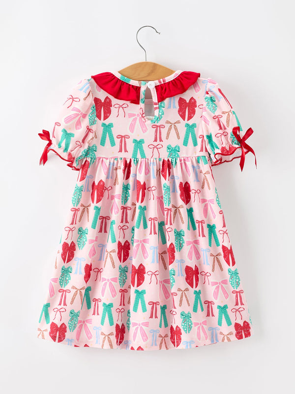 A short-sleeve baby dress with a pink background and an all-over bow print in red, pink, and teal, featuring a red ruffled collar and red tie details on the sleeves, shown hanging on a wooden hanger against a white background.