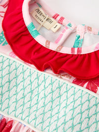 Close-up of a children's top with a bright red ruffled neckline, a pink and white patterned inner collar, and a white smocked section with mint green mermaid-style stitching.