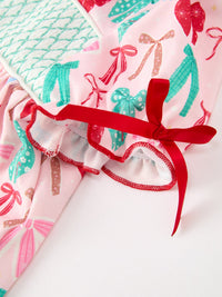 A close-up of a soft fabric product, likely a pillow or cushion cover, featuring a light pink background printed with colorful Christmas-themed silhouettes such as stockings, ornaments, and bows. The fabric is accented with a red ruffled trim and a red ribbon bow.