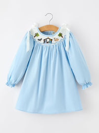 Light blue long sleeve girls dress with smocked neckline featuring embroidered nativity scene, palm trees, animals, and bow shoulder straps.