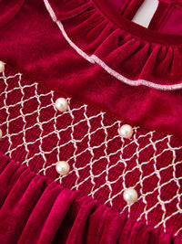 Burgundy velvet girls dress featuring white smocked waist detail, pearl accents, and delicate ruffle neckline for the holiday season.