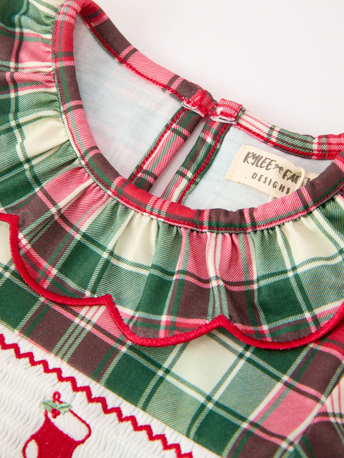 Green plaid girls Christmas dress with ruffle collar, red piping, and smocked chest panel featuring embroidered holiday stocking detail.